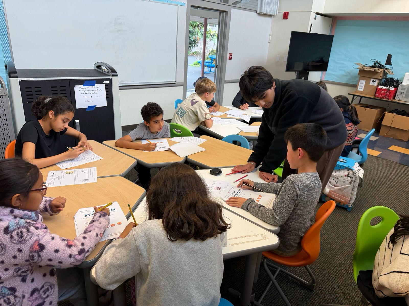 Devansh helping a group of younger students work through Math Kangaroo practice problems at a classroom table.